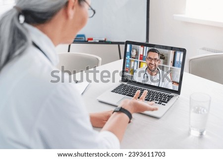 Adult female doctor has video call and chatting with colleague on laptop in clinic. Medical workers teamwork. Briefing about disease, illness treatment Royalty-Free Stock Photo #2393611703