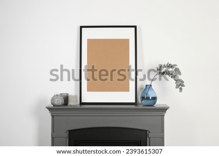 Empty frame, candles and eucalyptus branch in vase on fireplace near white wall indoors. Interior design