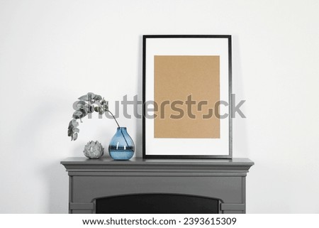 Empty frame and eucalyptus branch in vase on fireplace near white wall indoors. Interior design