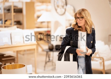 Mid aged woman standing in the interior design shop and looking around. Blond haired female wearing glasses and jacket. Royalty-Free Stock Photo #2393616361