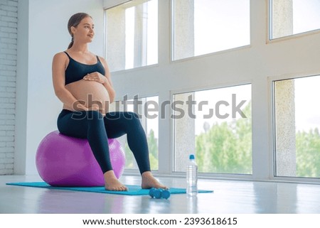 Happy pregnant woman sitting on a fitness ball and touching her belly with look in window. Caucasian expectant mother doing supportive physical exercises for smooth pregnancy Royalty-Free Stock Photo #2393618615
