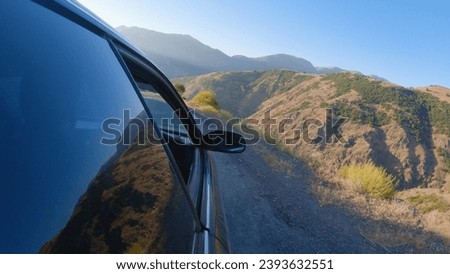 A car on a narrow mountain road. The mountains are reflected in the glass of the car. The camera is attached to the side of the car. In the background there is a beautiful view of the mountains