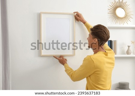 Young man hanging picture frame on white wall indoors, back view