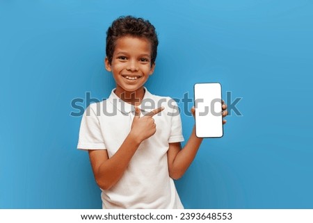 African-American boy in white polo shows blank smartphone screen on blue isolated background, 10-year-old child holds mobile phone and advertises copy space Royalty-Free Stock Photo #2393648553