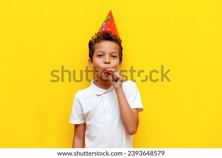 African American boy in festive hat celebrates his birthday and blows trumpet on blue isolated background, 10 year old child rejoices and makes noise for the holiday