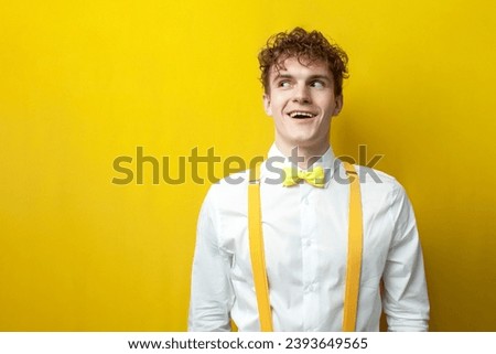 young funny guy in bow tie and suspenders looks in surprise to the side with his mouth open on yellow isolated background, nerdy student in festive outfit looks at copy space