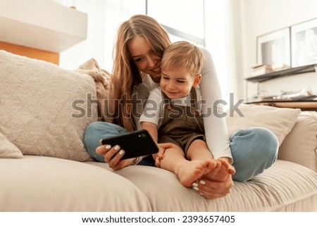 young mother and her little son are sitting on the sofa and using smartphone, 2-year-old boy is looking at his mobile phone and smiling with his parent, woman is watching cartoons online