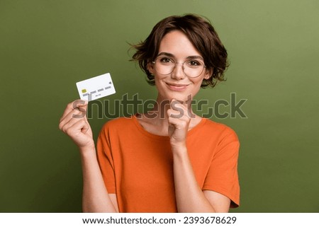 Photo of clever positive girl dressed orange t-shirt in glasses holding credit card arm on chin isolated on khaki color background Royalty-Free Stock Photo #2393678629