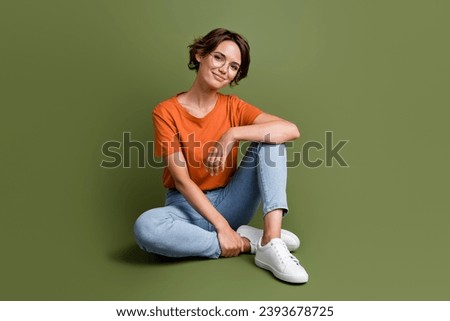 Full size photo of smart good mood woman dressed orange t-shirt jeans in glasses sit on floor smiling isolated on khaki color background