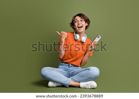 Full size photo of excited woman wear earphones sit look directing empty space smartphone in hand isolated on khaki color background