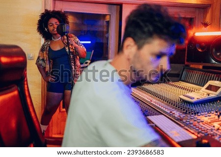 A young talented vocalist is standing in front of a microphone, creatively expressing while recording a part of the vocals for a song. With her is a sound engineer operating a large mixing console.