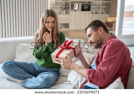 Pleasantly smiling merry European woman looking with excitement as intrigued interested man trying to unwrap giftbox with red ribbon. Unexpected surprise for birthday, anniversary, New Year Royalty-Free Stock Photo #2393699071