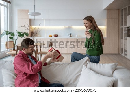 Happily laughing merry European woman looking with joy, fun and excitement as intrigued interested man looks at giftbox with red ribbon, wants to unwrap unexpected surprise for birthday, special event Royalty-Free Stock Photo #2393699073