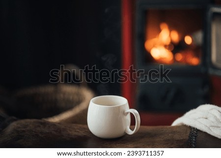 White cup with hot tea with burning fireplace on the background in cozy log cabin.