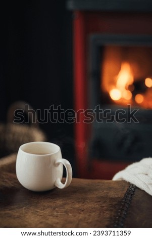 White cup with hot tea with burning fireplace on the background in cozy log cabin. Royalty-Free Stock Photo #2393711359