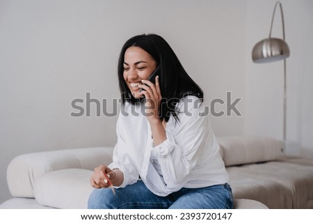 Laughing young African American woman in casual clothe sitting on cozy couch at home talking by phone smiling eyes closed. Funny conversation with friend, great joke. People candid emotions. Royalty-Free Stock Photo #2393720147