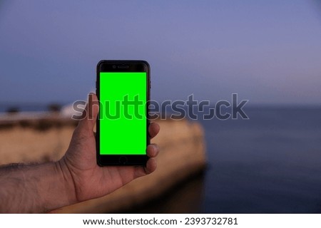 Male hand holding a smartphone with a green screen, and in the background the big cliff and the Chapel of Our Lady of the Rock in Porches, Algarve, Portugal