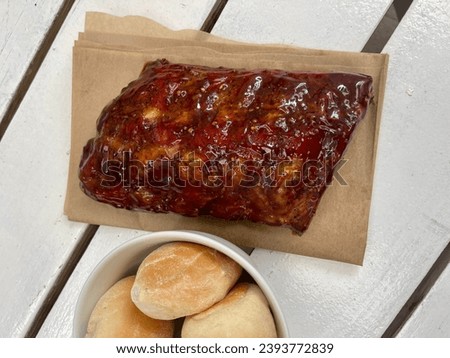 Overhead shot of a mouth-watering slab of smoked pork back ribs, smothered in a rich BBQ sauce, accompanied by a bowl of fresh buns on a white wooden table