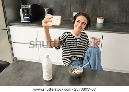 Stylish young woman, student taking selfie in the kitchen, drinking tea and eating breakfast, making pictures for social media app.