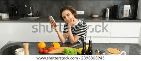 Portrait of cute brunette woman in the kitchen, cooking, searching recipe on smartphone, taking pictures for her food blog, cooking meal from vegetables, vegan salad.