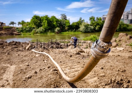 Long fire hose with metal coupling placed on river coast at the construction site for water supply, bridge construction is in progress. Royalty-Free Stock Photo #2393808303