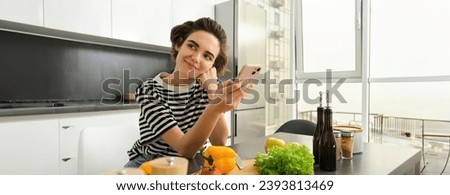 Portrait of brunette girl cooking food in the kitchen, searching recipes on social media app, holding mobile phone, standing near chopping board and vegetables, preparing healthy vegetarian meal.