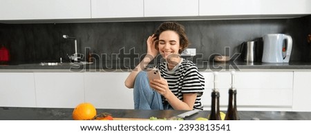 Portrait of young woman smiling, laughing and using smartphone while cooking, searching recipes on smartphone app, preparing meal from vegetables in her kitchen at home.