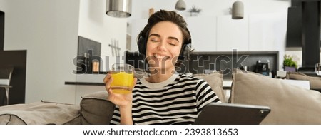 Close up portrait of smiling, happy brunette woman in living room, sitting on sofa and drinking orange juice, reading on digital tablet, using her gadget and resting at home.