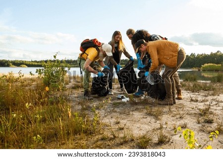 This image captures a group of individuals actively participating in a lakeside cleanup. Dressed in casual outdoor attire and armed with garbage bags and gloves, they are bent over, picking up litter