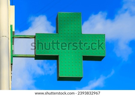 Green medical cross indicating the location of a pharmacy on a city street. Banner medical cross on a building against a background of blue sky and clouds. Pharmacy sign.