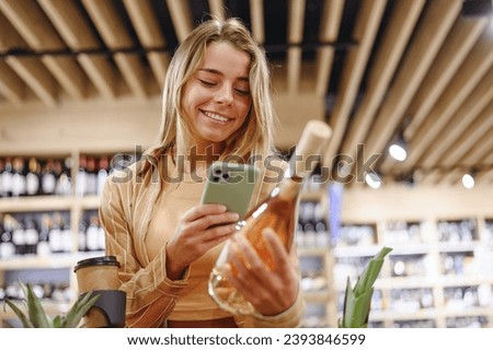 Young customer woman wearing casual clothes hold wine take photo on mobile cell phone shopping at supermaket store grocery shop with trolley cart buy choose products in hypermarket. Purchasing concept