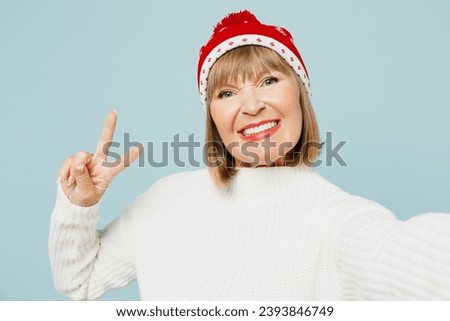 Close up merry elderly woman 50s years old wear sweater red hat posing doing selfie shot on mobile cell phone show v-sign isolated on plain blue background. Happy New Year celebration holiday concept
