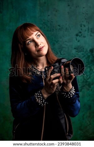 Portrait of adult woman photographer holds photo camera at green dark backdrop, pensive looking up away. Perfect photographer lady with camera, studio shot. Profession, occupation. Copy ad text space