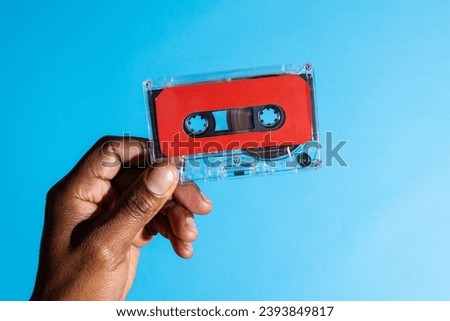 Close up of hand of african american man holding red cassette tape over blue background. Music, sound, listening, entertainment and nostalgia concept. Royalty-Free Stock Photo #2393849817
