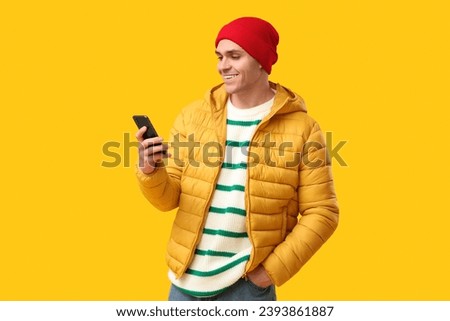 Young man in stylish puffer jacket and hat with mobile phone on yellow background