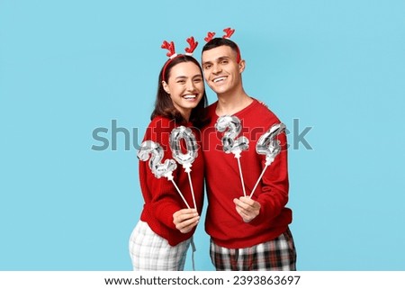 Happy young couple in Christmas pajamas and with balloons in shape of figure 2024 on blue background