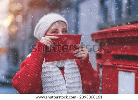cheerful woman in a red coat and white hat  putting a card in the red postbox and walking around an English city on a snowy day
