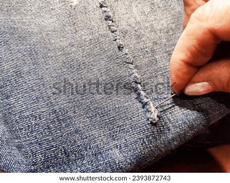 Woman repairing a pair of blue jeans. Fingers with a needle and torn jeans fabric. Sewing up a tear on blue fabric