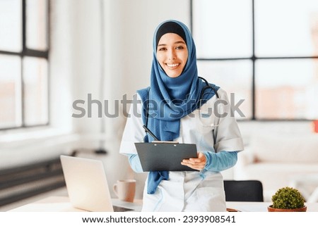 Islamic woman doctor in traditional religious hijab stands, smiles  in camera and takes notes
