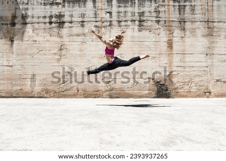 Side view of woman jumping in the air performing split over cement textured background on sunny day.
