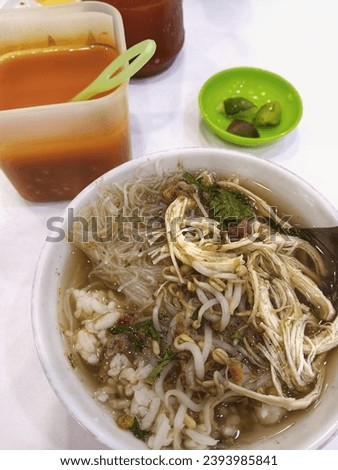 A bowl of soto containing rice, vermicelli, shredded chicken and bean sprouts doused in broth is served with chili sauce and lime slices on a white table. Royalty-Free Stock Photo #2393985841