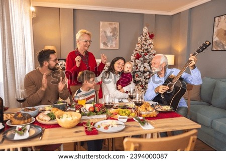 Happy multi-generation family gathered around the table playing the guitar and singing Christmas songs while having Christmas dinner all together at home Royalty-Free Stock Photo #2394000087
