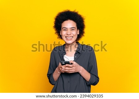 Positive joyful african american or brazilian curly haired young woman, using her smartphone, messaging with friends on social media, making online order, answering email, isolated yellow background