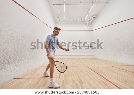 redhead sportsman in active wear holding squash ball and racquet while playing inside of court Royalty-Free Stock Photo #2394031505