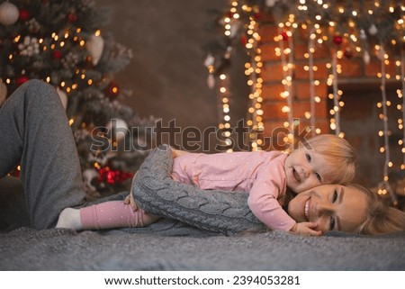 A blonde woman in a gray suit and her child in pink pajamas are lying on a bed near a Christmas tree. happy family time