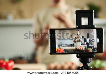 Closeup of smartphone filming young middle eastern blogger tossing up tomato while standing near kitchen table, focus on smartphone, copy space Royalty-Free Stock Photo #2394056055