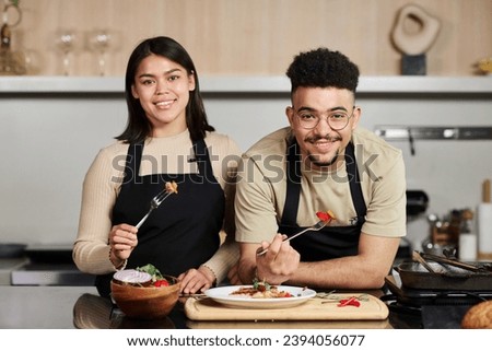 Medium shot of smiling young middle eastern man and happy hispanic woman standing by kitchen counter holding forks with food bites