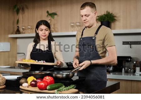 Medium shot of young hispanic woman and caucasian chef standing by kitchen stove about to fry chopped vegetables in pan