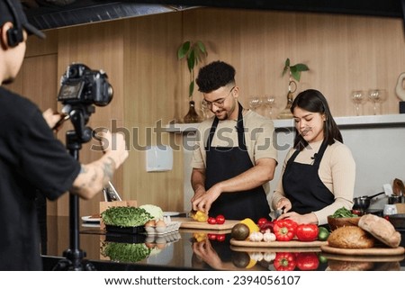 Medium shot of young middle eastern guy and hispanic girl standing by kitchen counter cutting vegetables while being filmed by cameraman