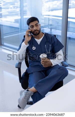 Biracial male doctor talking on smartphone and holding cup of coffee in hospital office. Medicine, healthcare, communication and medical services, unaltered.